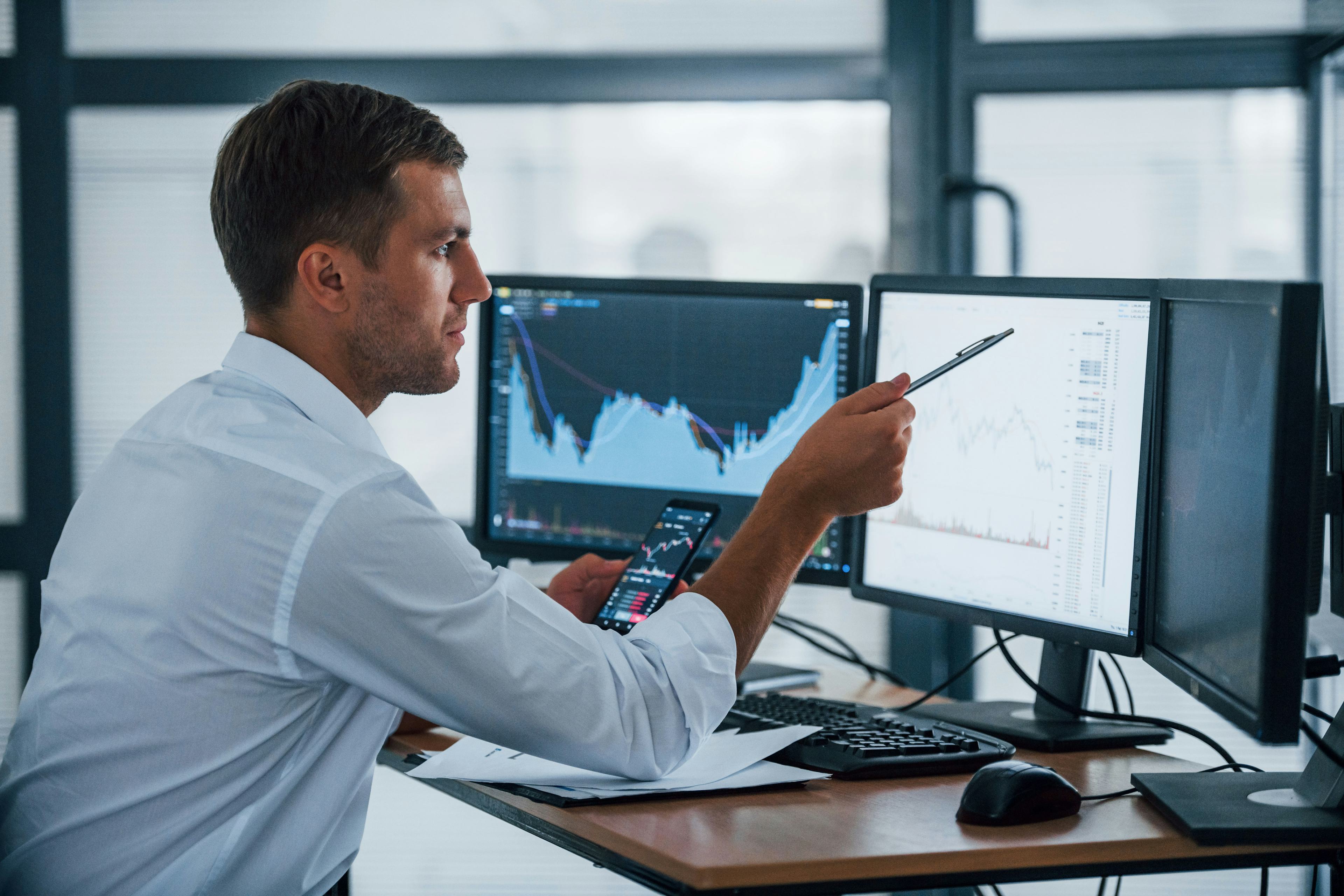 Young businessman in formal clothes is in office with multiple screens. Conception of exchange and money.