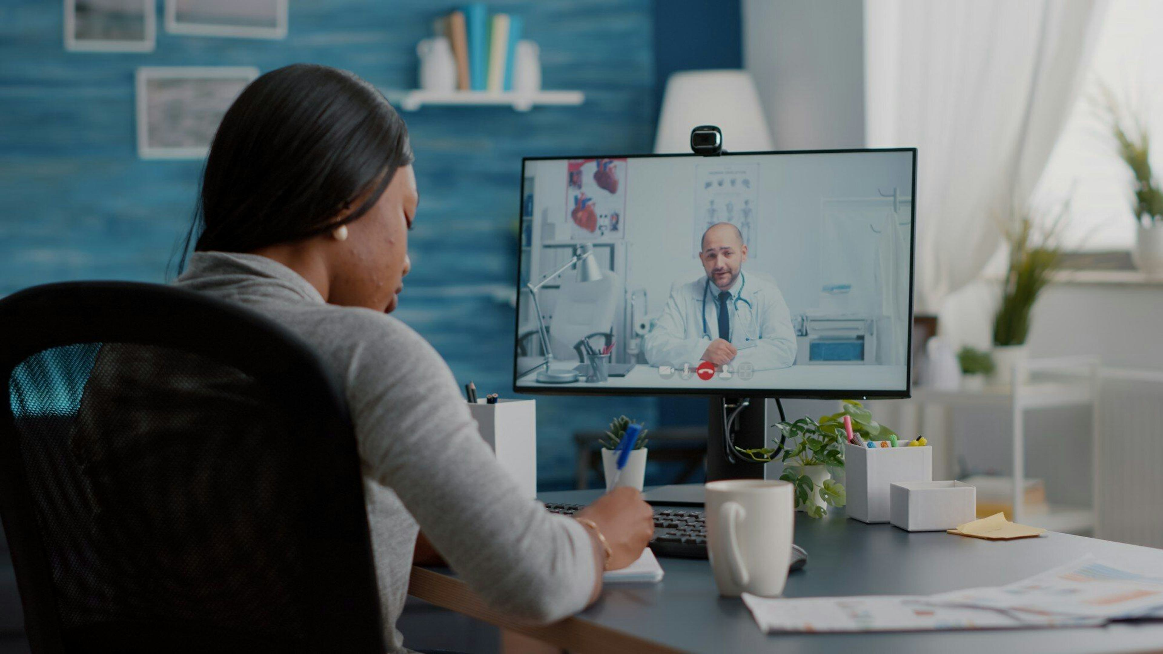 young woman writing in notebook as doctor speaks to her through video call screen