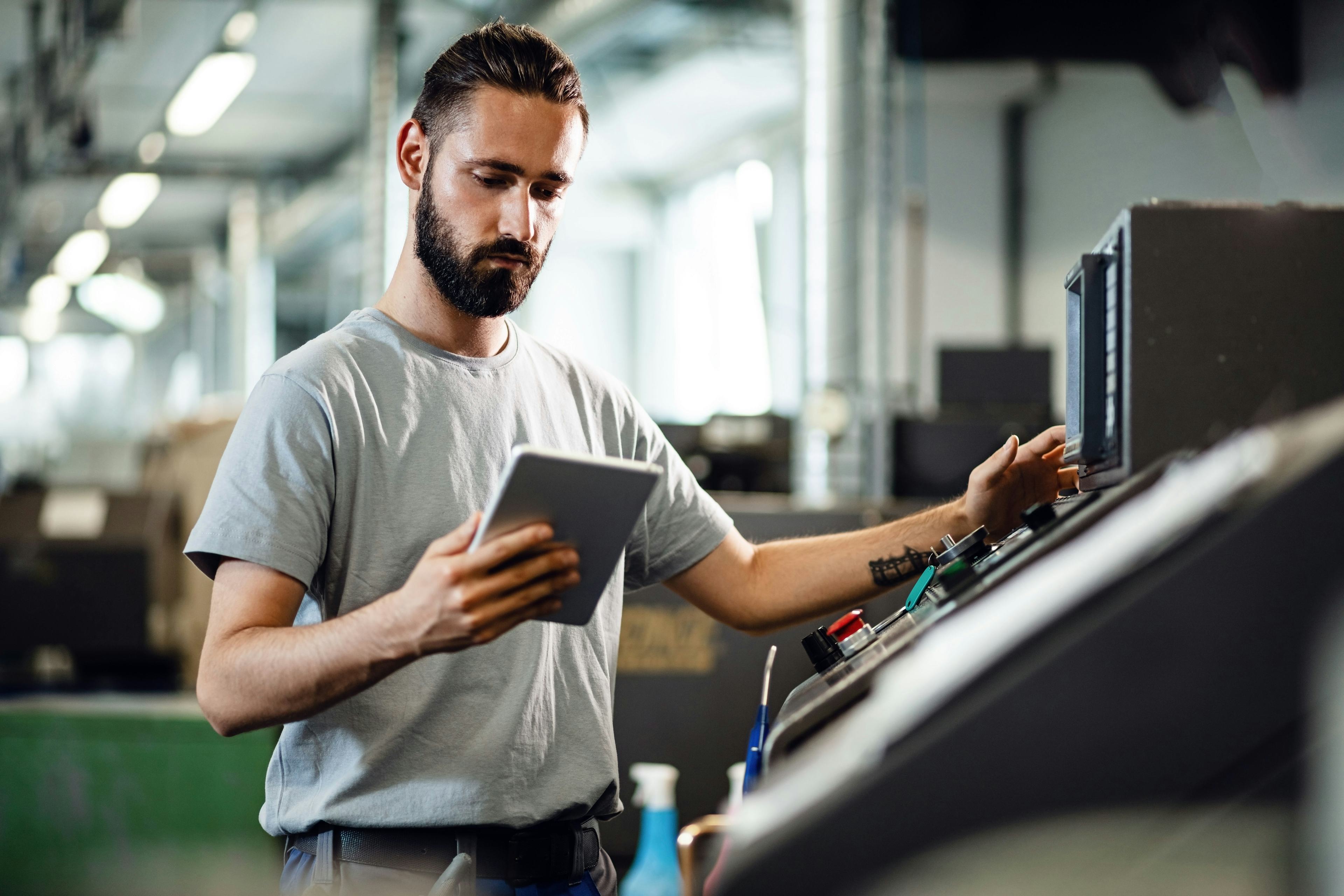 Young worker operating a CNC machine while using digital tablet in industrial facility