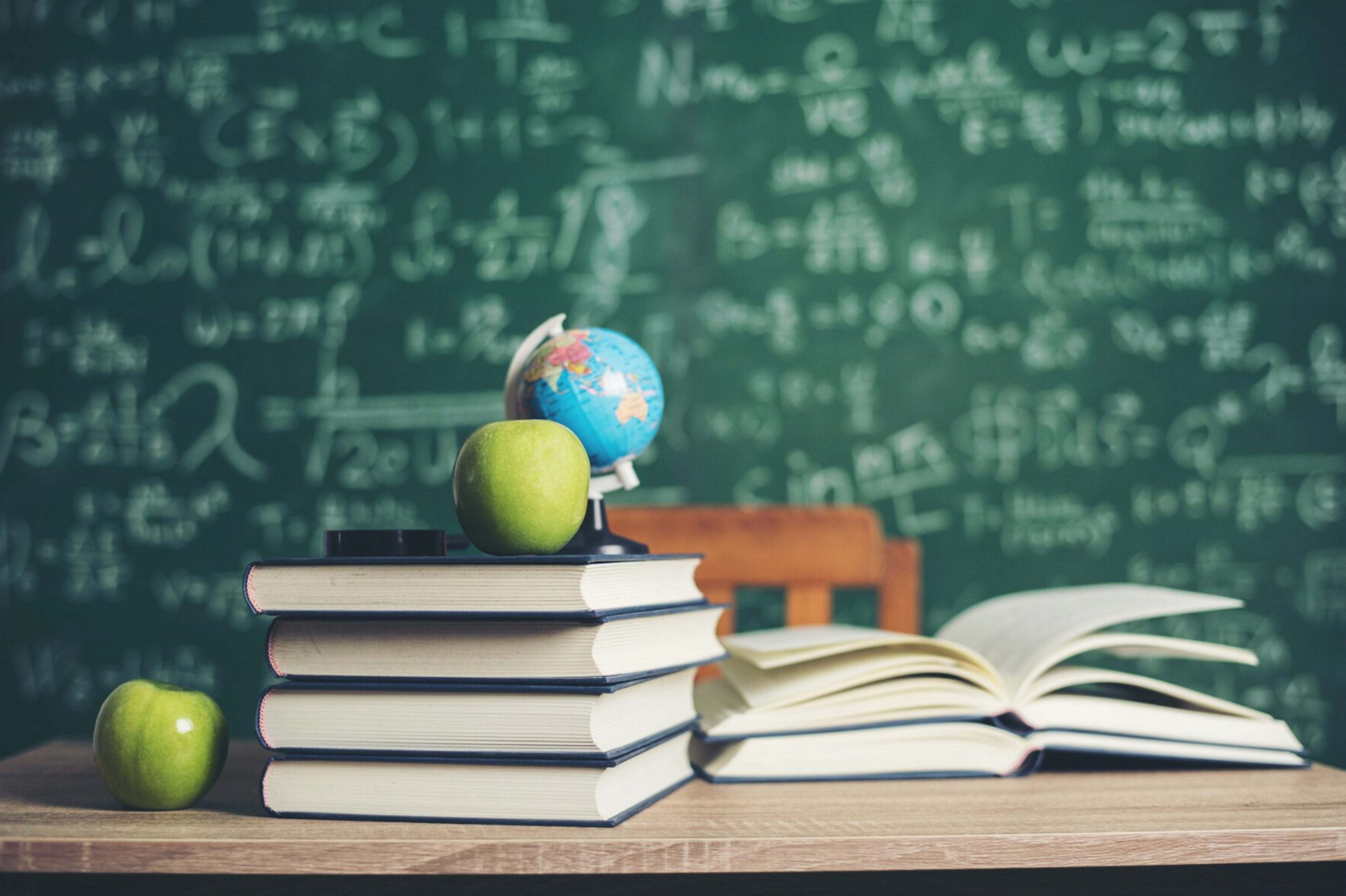 stack of books with green apples and globe and blackboard lesson in background