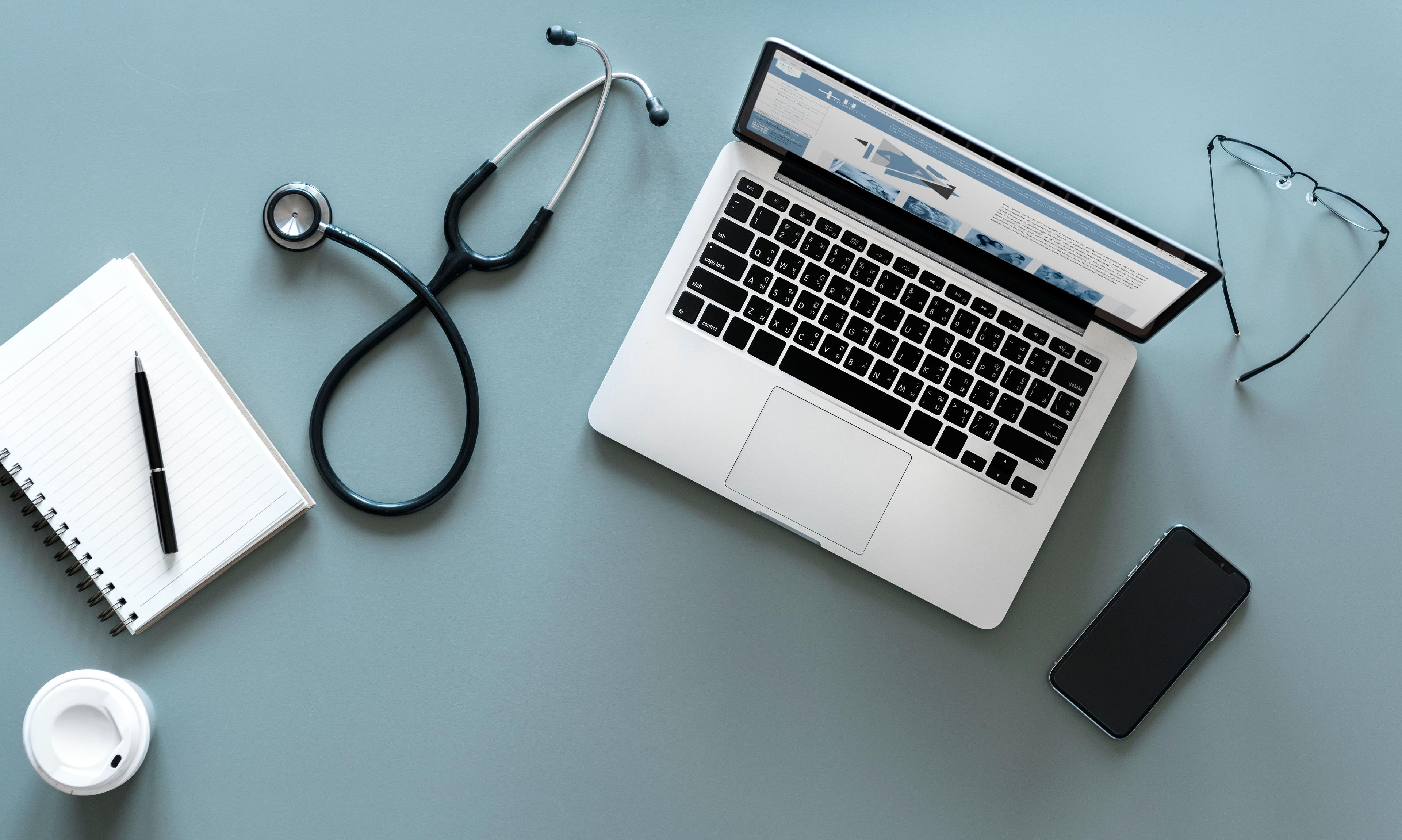 aerial shot of notebook, coffee cup, phone, glasses, and stethoscope around a laptop