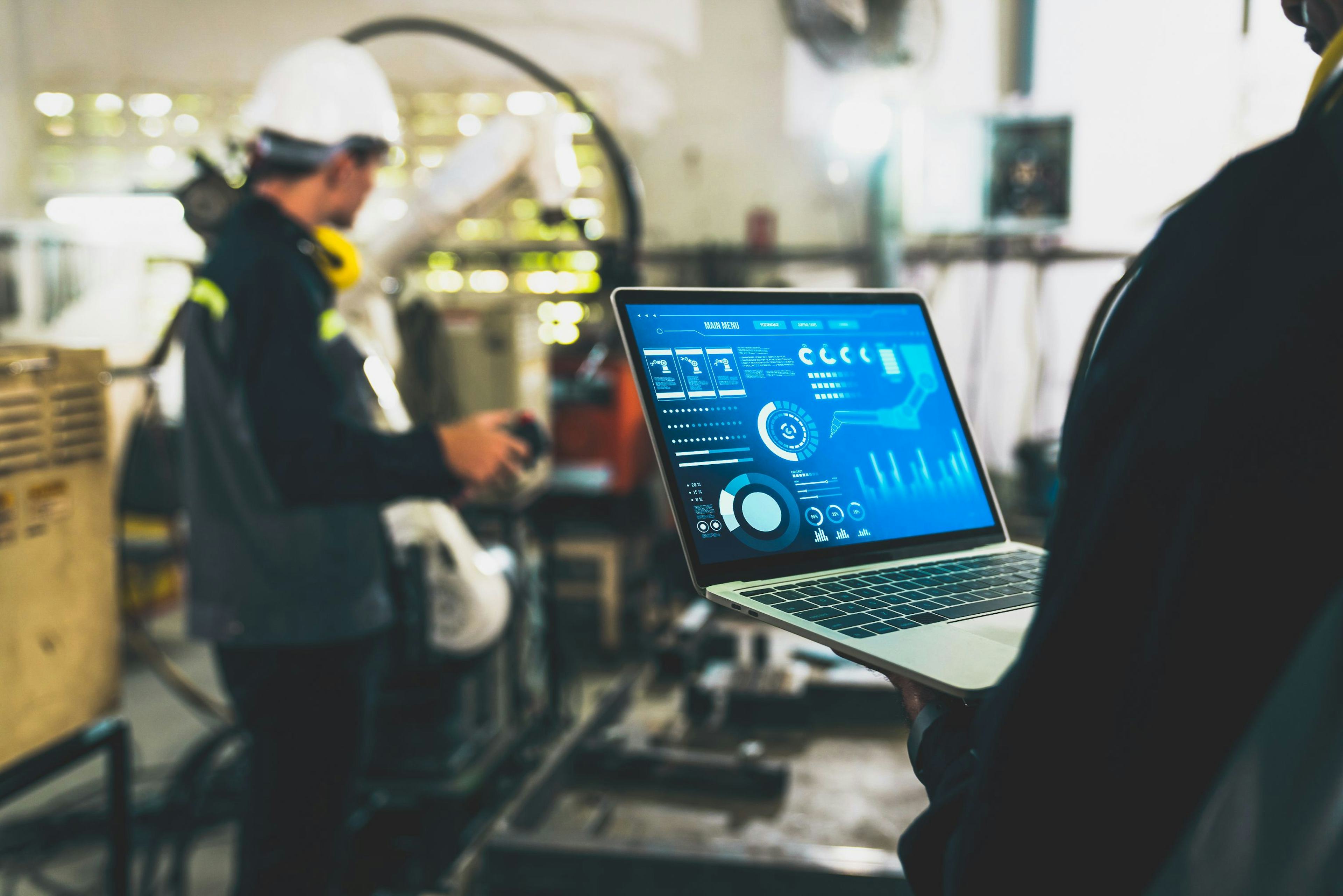 Factory workers working with adept robotic arm in a workshop with graphical data displaying on a tablet