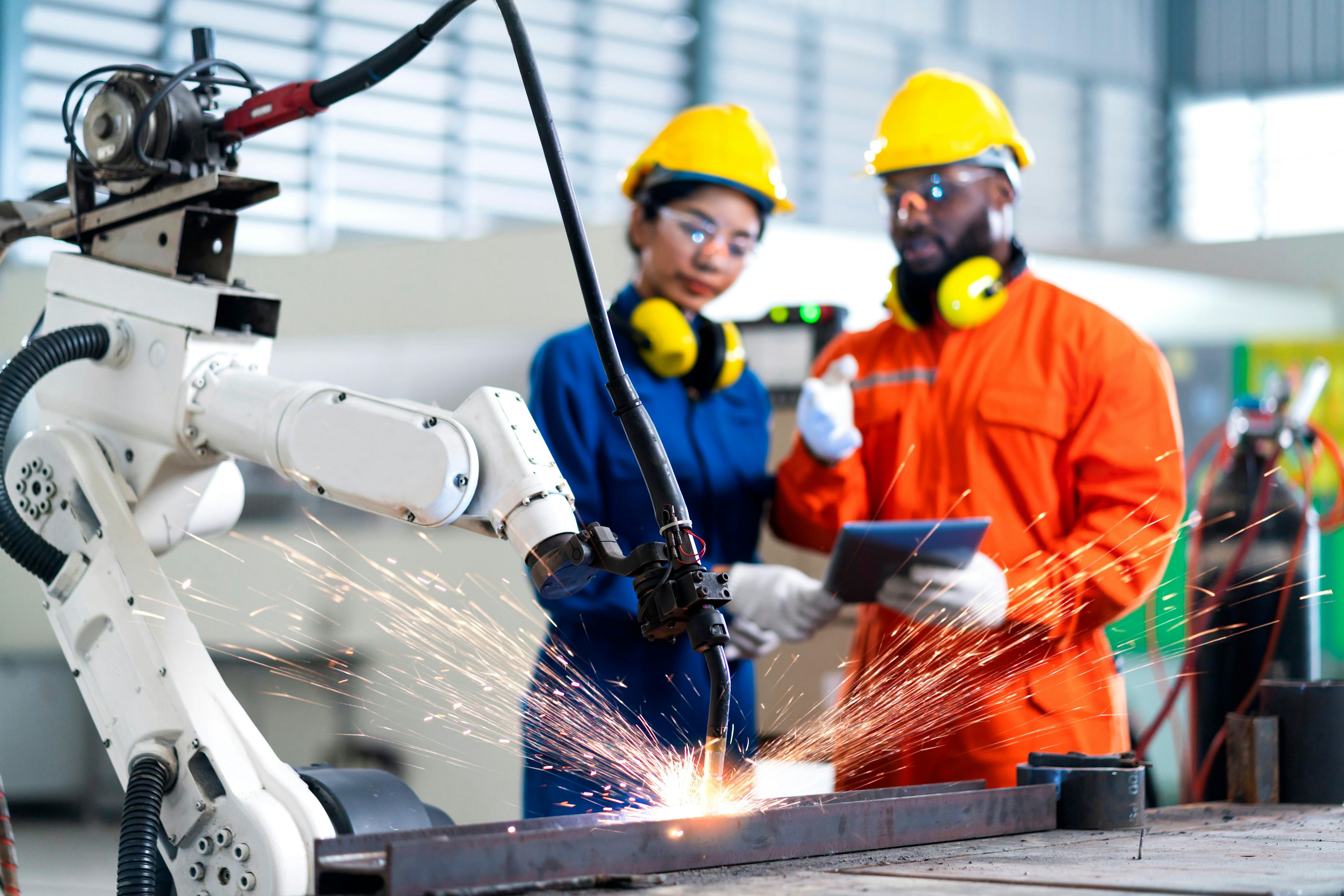 Robotic arm producing sparks while manufacturing products while two people in hardhats and goggles stand in the backdrop