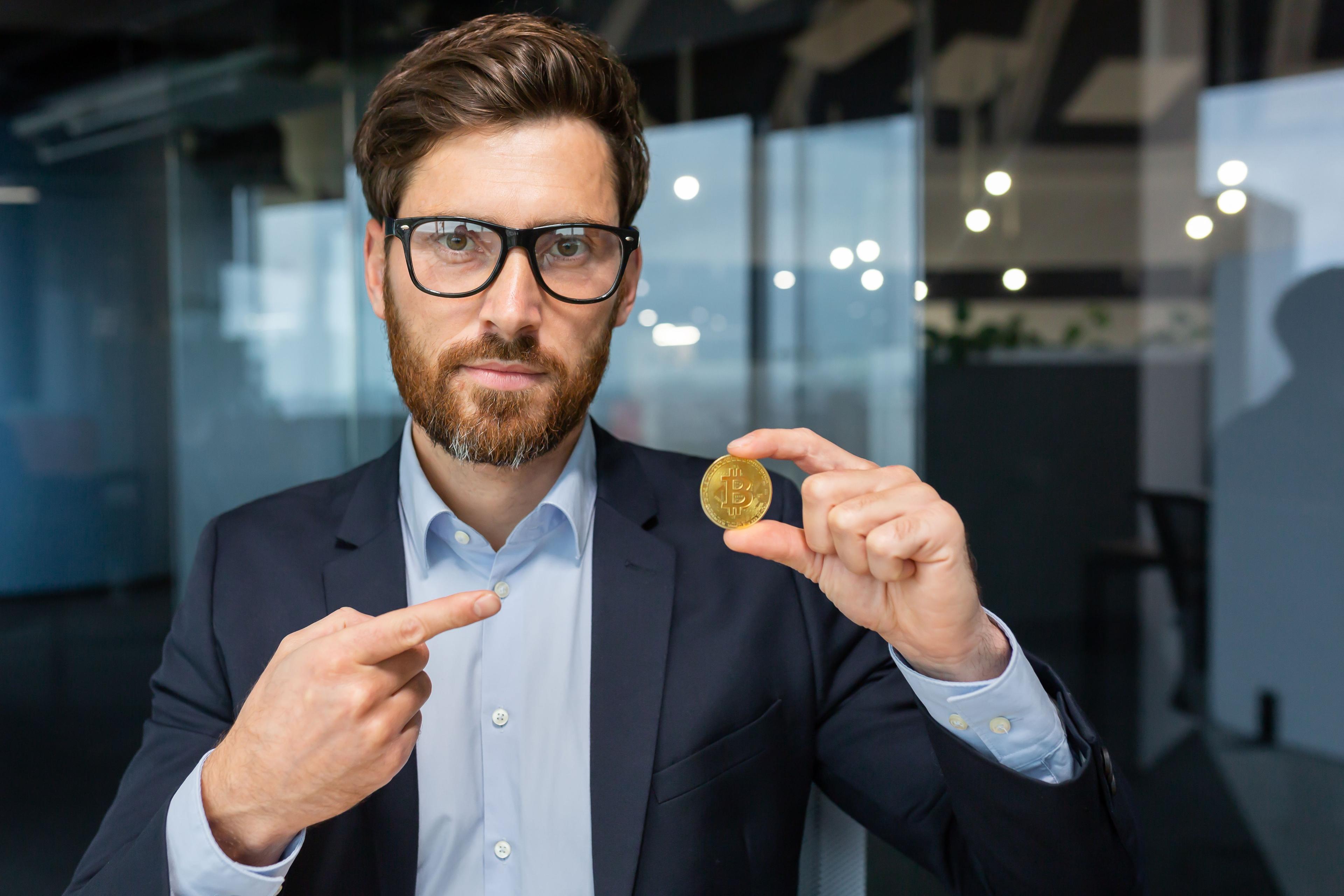 professional businessman in a dark navy blue suit and blue shirt holding a physical bitcoin