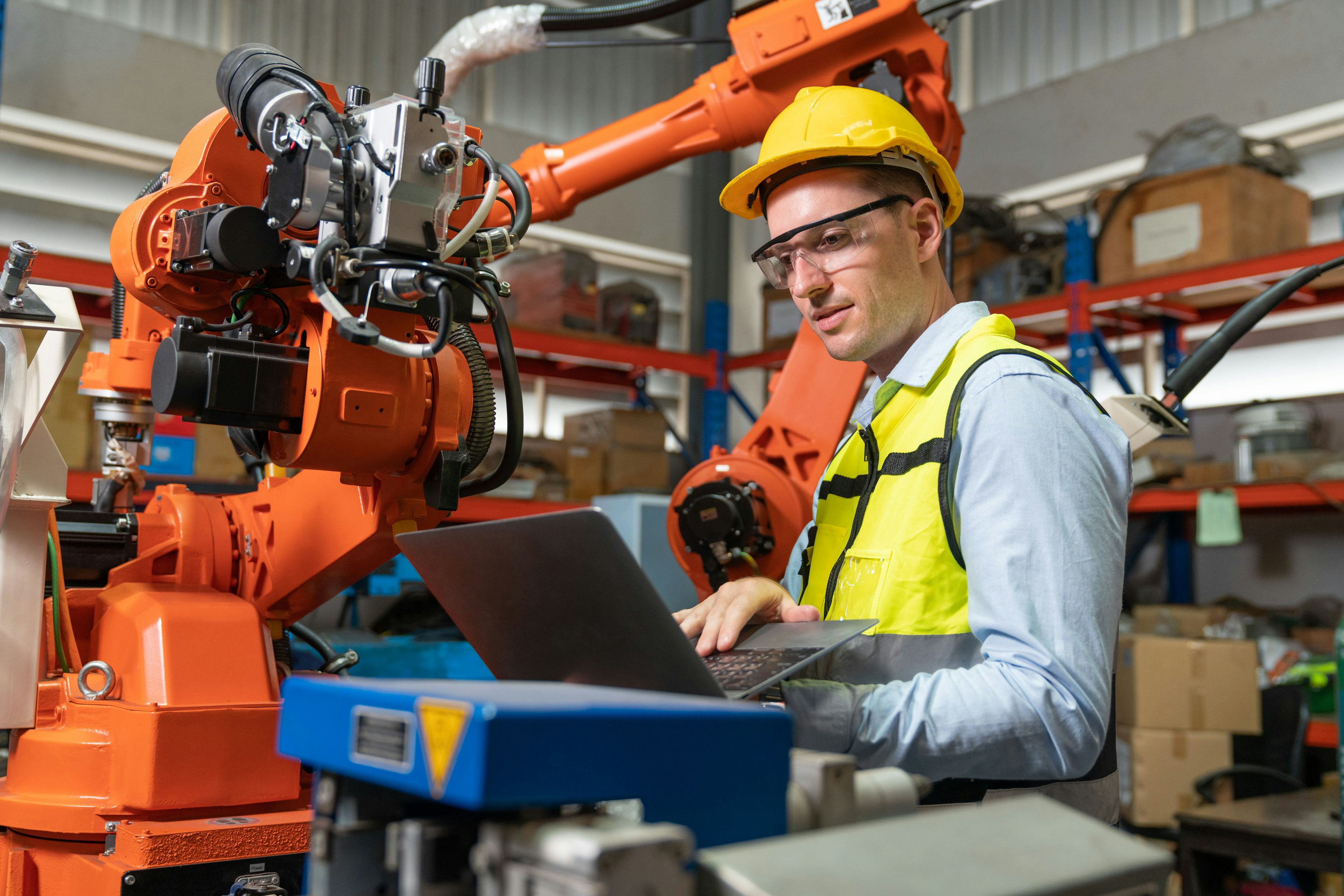 industrial worker looking at laptop while standing by robotic arm