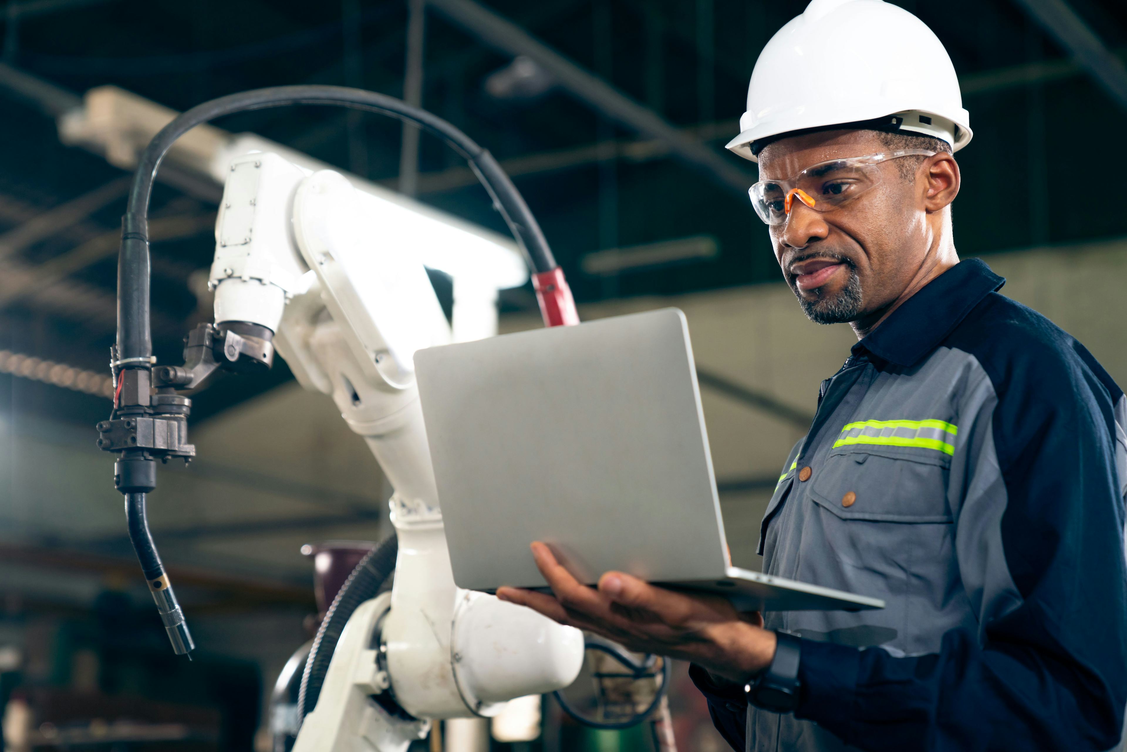 working man wearing hard hat in industrial setting with robotic arm behind him