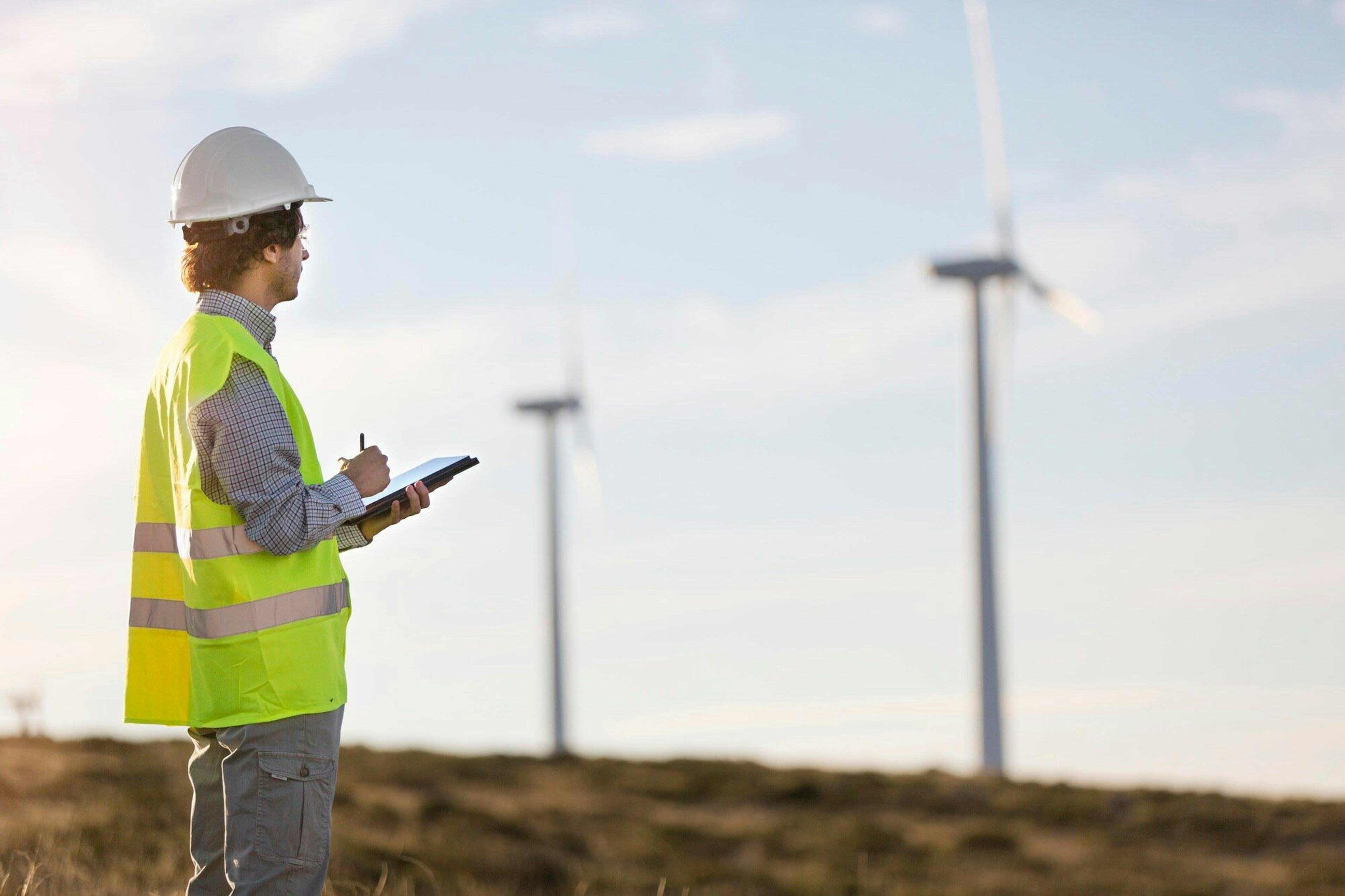 man wearing fluorescent yellow suit with industrial hard hat on as he holds tablet and oversee wind turbines